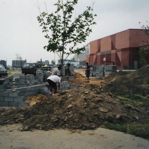 Masons laying concrete block for the flower boxes during construction, with excavated ground and stacked block across the site